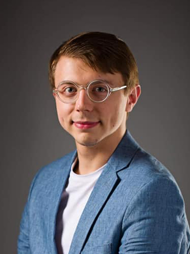 Treyton Blackburn color photo, looking at camera, wearing blue suit jacket and round rimmed glasses