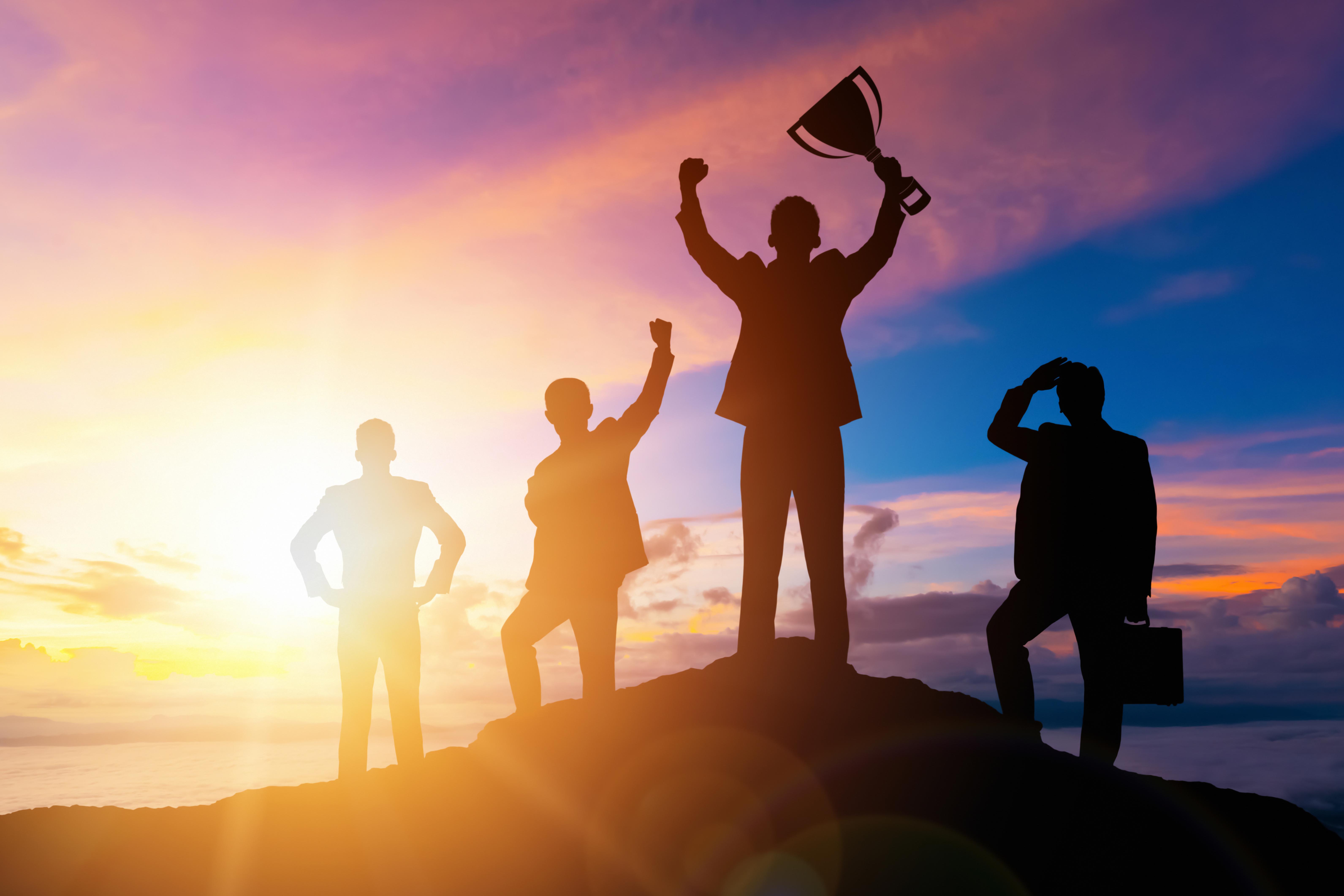 four individuals in front of a sunset mountain scape. One is triumphantly lifting a trophy.